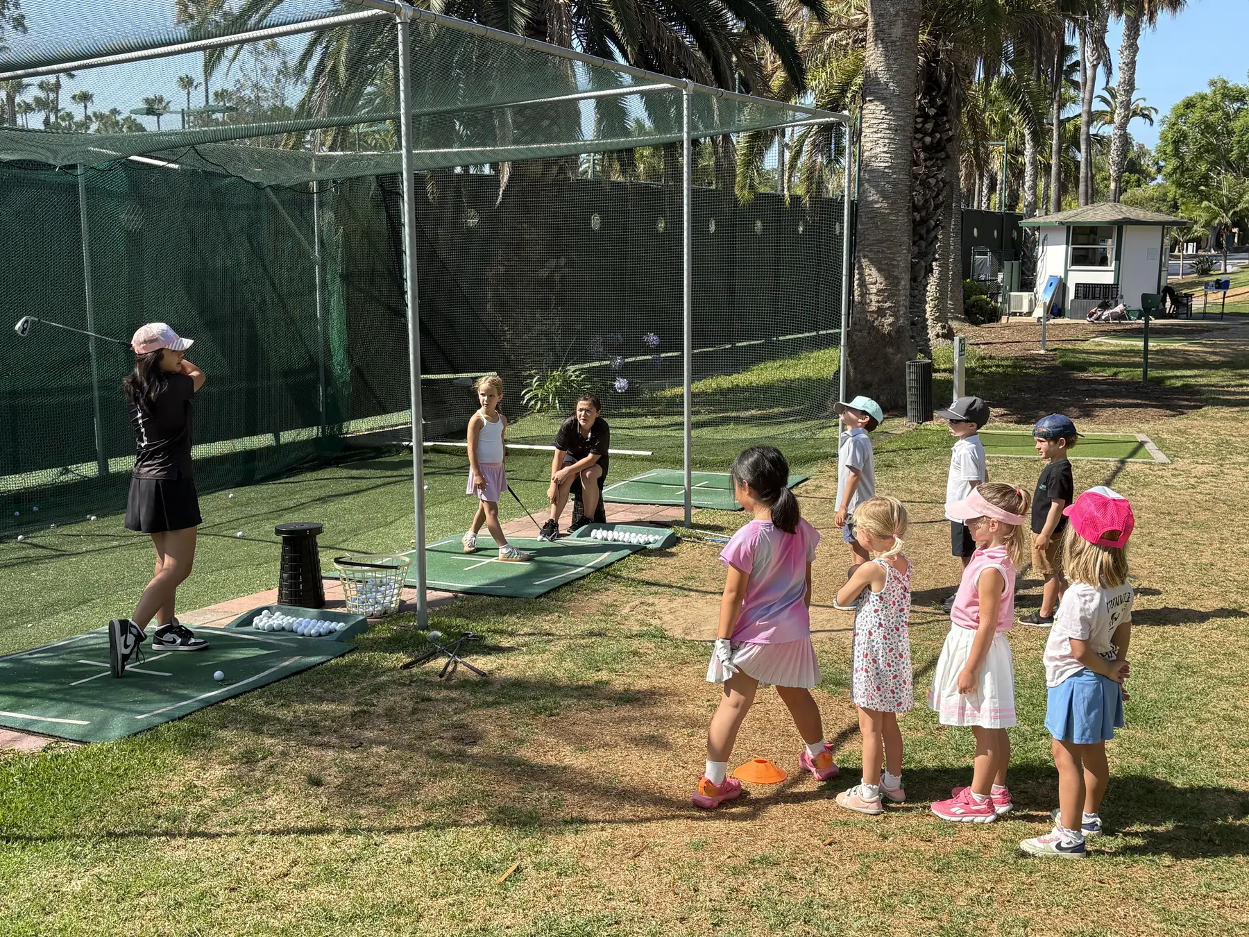Tiny Tees players lined up on the grass watching a coach swing at the practice nets